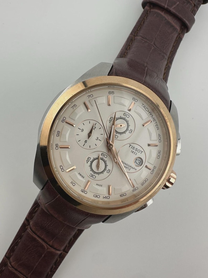 May include: A Tissot chronograph quartz watch with a silver face, gold bezel, and brown leather strap. The watch face displays multiple sub-dials and the date. The watch is angled, showcasing the details of the dial and strap.