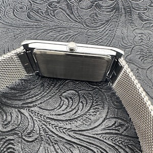 May include: A rectangular silver wristwatch with a mesh band. The watch face is not visible. The watch is set against a dark, textured background with a floral pattern. The watch band appears to be made of woven metal.