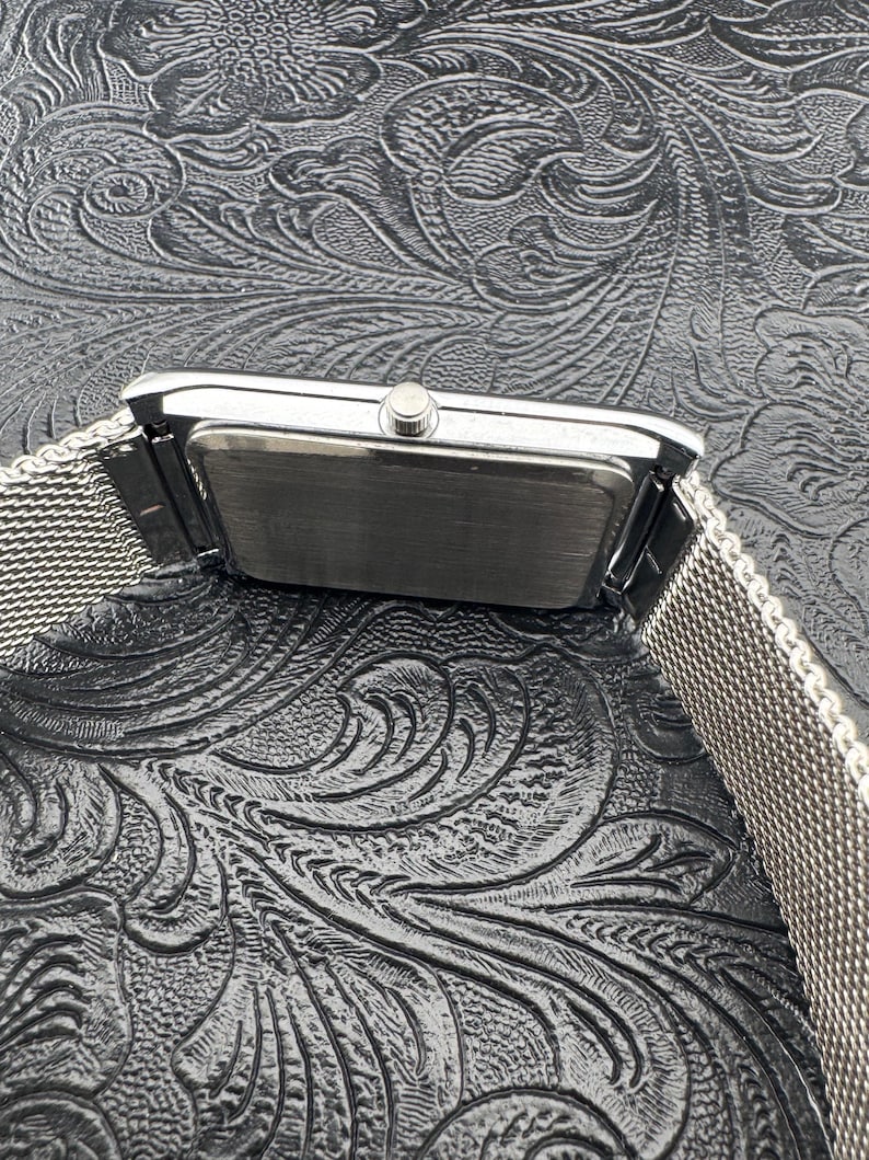 May include: A rectangular silver wristwatch with a mesh band. The watch face is not visible. The watch is set against a dark, textured background with a floral pattern. The watch band appears to be made of woven metal.
