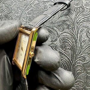 May include: A gold Seiko Quartz watch with a rectangular face and black leather strap. The watch face is cream-coloured with Roman numerals and gold hands. The watch is held by a gloved hand.