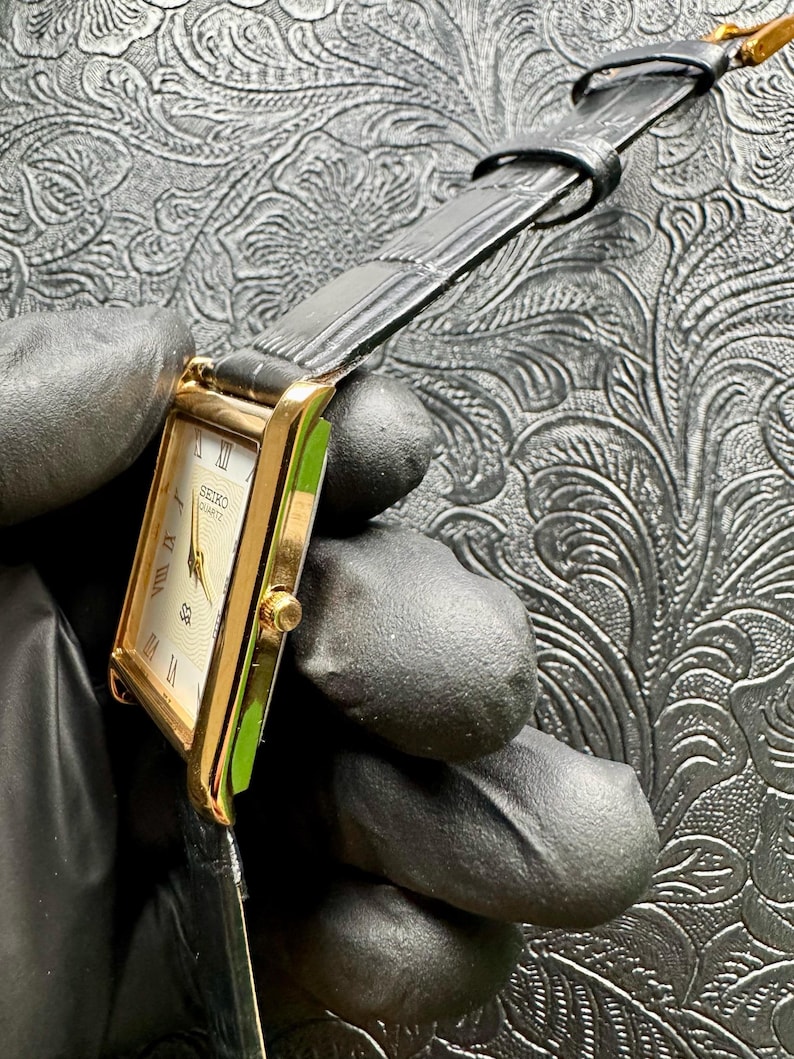 May include: A gold Seiko Quartz watch with a rectangular face and black leather strap. The watch face is cream-coloured with Roman numerals and gold hands. The watch is held by a gloved hand.