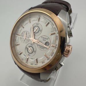 May include: A Tissot chronograph watch with a silver and gold-tone case and a brown leather strap. The watch face is white with three sub-dials and gold-tone hands. The watch has a tachymeter scale and the date displayed at the 3 o'clock position.
