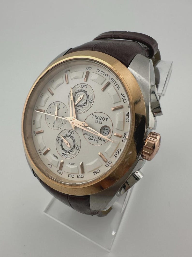 May include: A Tissot chronograph watch with a silver and gold-tone case and a brown leather strap. The watch face is white with three sub-dials and gold-tone hands. The watch has a tachymeter scale and the date displayed at the 3 o'clock position.
