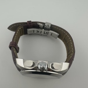 May include: A silver-toned wristwatch with a brown leather band. The watch face is round, and the band is secured with a silver clasp. The watch has two buttons on the side and a small crown.