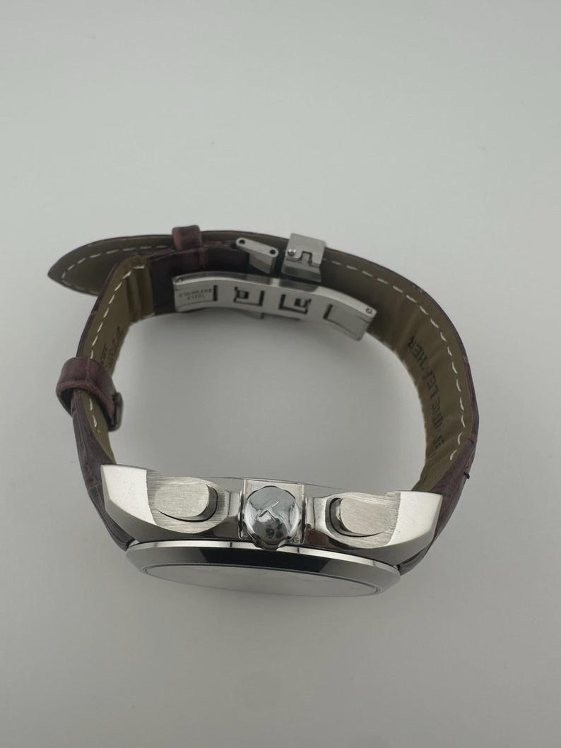 May include: A silver-toned wristwatch with a brown leather band. The watch face is round, and the band is secured with a silver clasp. The watch has two buttons on the side and a small crown.