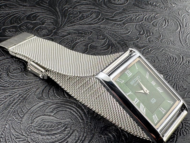 May include: A rectangular Seiko Quartz watch with a silver-tone case and a mesh band. The watch face is green with silver hands and Roman numeral hour markers. The watch is displayed on a textured black surface.