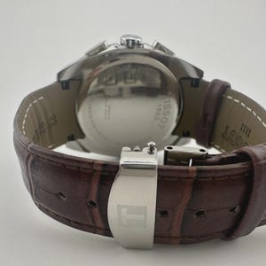 May include: A stainless steel Tissot watch with a brown leather strap. The watch features a silver clasp and a round face. The back of the watch has the text "Tissot 1853". The strap has a crocodile pattern.