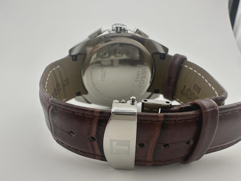 May include: A stainless steel Tissot watch with a brown leather strap. The watch features a silver clasp and a round face. The back of the watch has the text "Tissot 1853". The strap has a crocodile pattern.