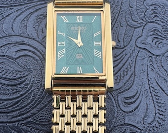 May include: A gold-toned rectangular watch with a green face and Roman numeral hour markers. The watch band is a matching gold-toned mesh style. The watch face has the brand name "SEIKO QUARTZ" printed on it.