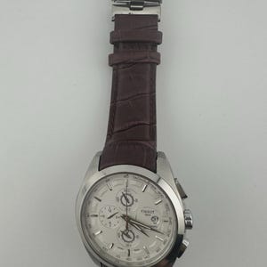 May include: A silver-toned Tissot chronograph watch with a white face and brown leather strap. The watch features multiple sub-dials, a date window, and a tachymeter scale. The strap has a crocodile pattern.