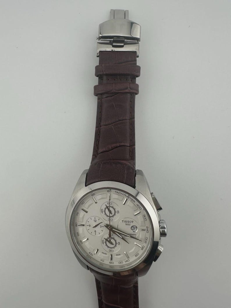 May include: A silver-toned Tissot chronograph watch with a white face and brown leather strap. The watch features multiple sub-dials, a date window, and a tachymeter scale. The strap has a crocodile pattern.
