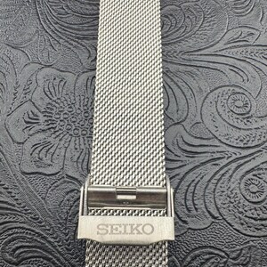 May include: A close-up of a silver-toned watch band with a mesh design. The band is secured with a clasp that has the brand name "SEIKO" engraved on it. The background is a dark, textured surface.