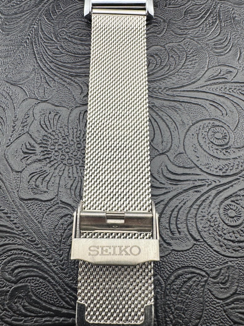 May include: A close-up of a silver-toned watch band with a mesh design. The band is secured with a clasp that has the brand name "SEIKO" engraved on it. The background is a dark, textured surface.