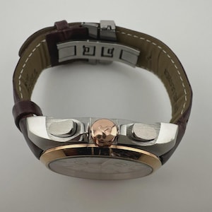 May include: A luxury wristwatch with a brown leather band and a silver and rose gold case. The watch features a round face with a detailed dial and two buttons on the side. The band is secured with a silver clasp.
