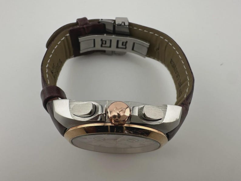 May include: A luxury wristwatch with a brown leather band and a silver and rose gold case. The watch features a round face with a detailed dial and two buttons on the side. The band is secured with a silver clasp.