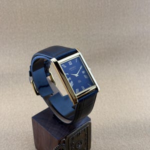 May include: A rectangular Seiko quartz wristwatch with a gold-tone case and a dark blue dial featuring Roman numeral hour markers. The watch has a dark blue leather strap with a textured finish and a gold-tone buckle closure.