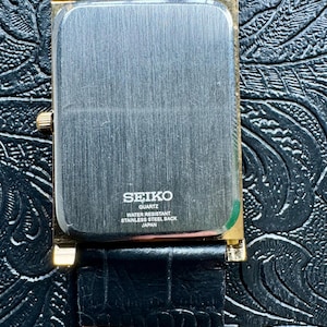 May include: A rectangular Seiko Quartz watch with a gold-toned case and a black leather strap. The watch face is silver with the Seiko logo and text indicating it is water-resistant and has a stainless steel back. The strap is textured.