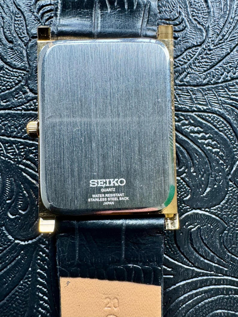 May include: A rectangular Seiko Quartz watch with a gold-toned case and a black leather strap. The watch face is silver with the Seiko logo and text indicating it is water-resistant and has a stainless steel back. The strap is textured.