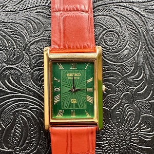 May include: A rectangular gold-toned Seiko Quartz watch with a green face and red leather strap. The watch face features Roman numerals and the Seiko logo. The strap has a textured, embossed pattern.