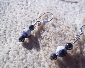 May include: A pair of dangle earrings featuring a combination of dark blue and white speckled beads and smaller, dark blue beads. The earrings have silver-colored ear wires and are displayed on a textured, off-white surface.