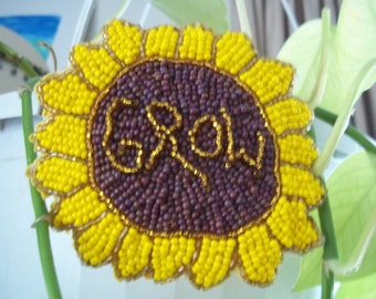 May include: A beaded sunflower with a brown center and yellow petals. The center of the sunflower has the word "GROW" spelled out in gold beads.