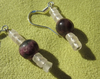 May include: A pair of handmade earrings featuring a dark purple, round bead and clear, cylindrical beads. The earrings have silver-colored ear wires. The jewelry is displayed on a light green surface.