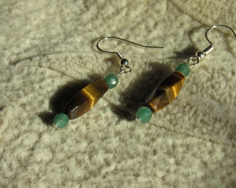 May include: A pair of dangle earrings featuring tiger's eye and green beads. The earrings have a silver hook and are set against a textured, off-white background. The tiger's eye beads are brown with golden stripes.