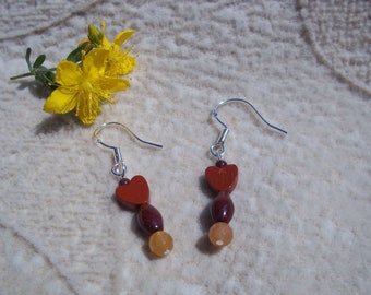 May include: A pair of dangle earrings with silver hooks. Each earring features a red heart-shaped bead, a dark red oval bead, and a light orange round bead. A yellow flower with green leaves is in the background.