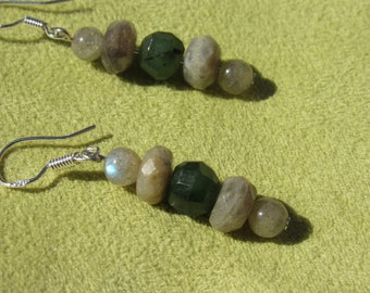 May include: A pair of earrings with silver hooks. Each earring features a series of beads in shades of gray, tan, and green. The beads are various shapes, including round and faceted. The earrings are displayed on a light green surface.