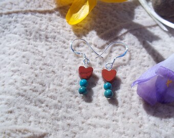 May include: Silver dangle earrings featuring a red heart-shaped stone and two turquoise beads. The earrings are displayed on a textured white surface, with yellow and purple flowers in the background.