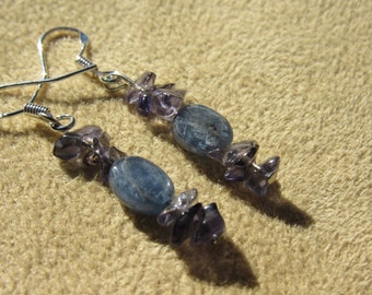 May include: A pair of dangle earrings featuring blue oval-shaped stones and small, irregular purple stones. The earrings have silver-colored hooks. The background is a tan, textured surface.