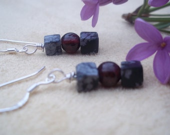 May include: A pair of handcrafted earrings with silver hooks. Each earring features a dark red bead between two square, speckled gray and black beads. The earrings are displayed on a cream-colored surface, with purple flowers in the background.