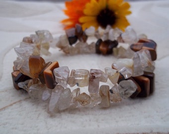 May include: A gemstone bracelet featuring a mix of clear, brown, and amber-colored stones. The bracelet is made of irregular-shaped stones and is displayed on a white surface. A yellow flower with an orange center is in the background.