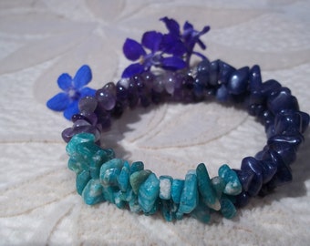 May include: A multi-colored gemstone bracelet with blue, purple, and green stones. The bracelet is made of small, irregular shaped stones and is accented with blue flowers.
