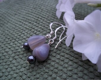 May include: A pair of dangle earrings featuring gray and white agate stones and small, dark gray beads. The earrings have silver-colored hooks. The background is a textured green surface with white flowers.