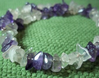 May include: A bracelet made of polished, irregular-shaped amethyst and clear quartz stones. The stones are strung together on a flexible cord, creating a piece of jewelry with a natural, earthy aesthetic. The bracelet is shown against a green background.