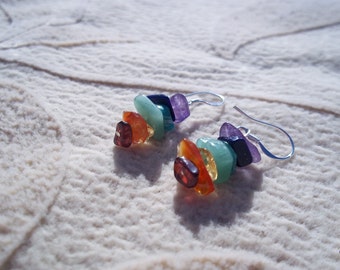 May include: A pair of silver earrings with a stack of colorful gemstones. The stones are arranged in a rainbow pattern, starting with purple, then blue, green, yellow, orange, and red.