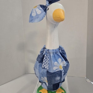 May include: A whimsical goose figurine with a white body, yellow beak and feet, and a denim outfit. The goose wears a blue denim bonnet with daisy accents and a matching skirt. The base is a green oval.