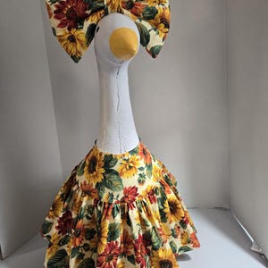 May include: A white goose figurine wearing a gathered dress and a matching oversized bow, both featuring a vibrant sunflower print with yellow, orange, and dark red petals against a cream background, with green leaves interspersed throughout.