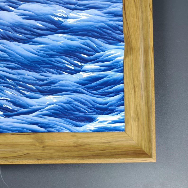 May include: A framed art print featuring an abstract depiction of ocean waves in shades of blue and white. The artwork is encased in a natural wood frame, showcasing the texture and grain of the wood.