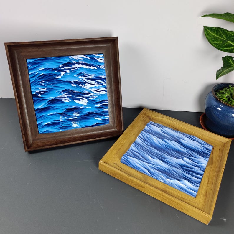 May include: Two framed art pieces with a blue and white wave design. One frame is dark brown, the other is light brown. The artwork depicts a textured ocean scene. The frames are square and sit on a dark surface.