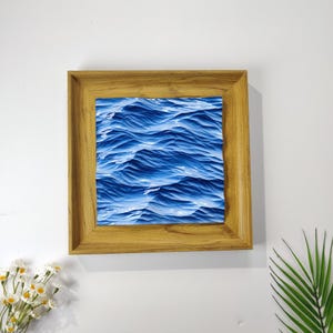 May include: A framed artwork featuring a close-up of blue ocean waves. The artwork is encased in a light brown wooden frame. The waves are depicted in varying shades of blue, creating a textured, dynamic effect. The overall theme is nautical and coastal.