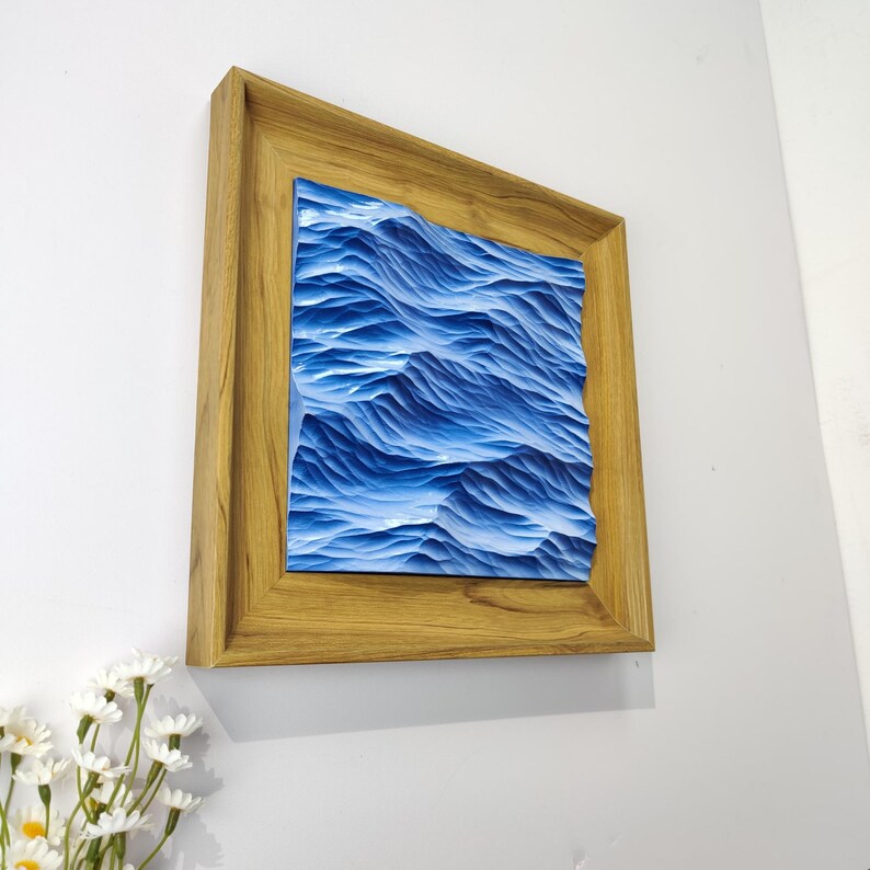 May include: A framed artwork depicting a textured blue ocean scene. The artwork is encased in a light brown wooden frame and mounted on a white wall. The ocean waves are a deep blue color.