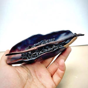 May include: A handcrafted wooden tea leaf holder with a dark purple interior and a natural wood exterior. The holder is filled with dark tea leaves and is held in a hand.