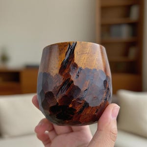 Op de afbeelding: Een handgesneden houten beker met een uniek, getextureerd oppervlak. De beker heeft een rijke bruine kleur met donkere accenten, die de natuurlijke nerf van het hout laten zien. De beker heeft een ronde, steelvrije vorm.
