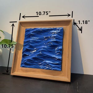 May include: Square wooden frame with a blue wave design, measuring 10.75 inches by 10.75 inches. The frame is 1.18 inches thick, showcasing a textured, ocean-themed artwork. The artwork is a vibrant blue, creating a sense of depth and movement.