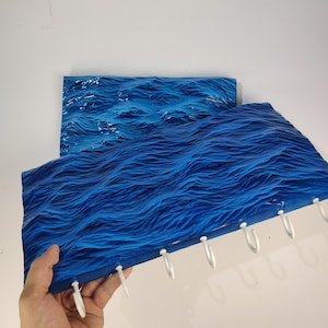 Può includere: Due pezzi di arte murale blu con texture a onde. Un pezzo è tenuto da una mano, rivelando ganci bianchi lungo il bordo inferiore. L'arte presenta diverse tonalità di blu, creando un realistico effetto onda oceanica. Il secondo pezzo è sullo sfondo.