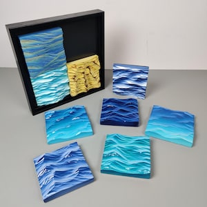 May include: A collection of ocean-themed art pieces. A framed artwork features blue and yellow textured sections. Several square blocks depict ocean waves in shades of blue and turquoise. The art pieces are displayed on a grey surface.