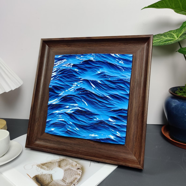 May include: A framed artwork depicting a close-up of ocean waves in shades of blue and white. The artwork is encased in a square, dark brown wooden frame, creating a contrast between the natural scene and the structured frame.