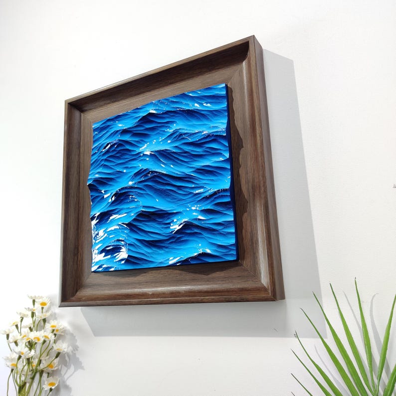 May include: A framed artwork depicting a stylized ocean scene. The artwork features a textured, three-dimensional effect with shades of blue and white, framed in a dark brown wooden frame. The piece is mounted on a white wall.
